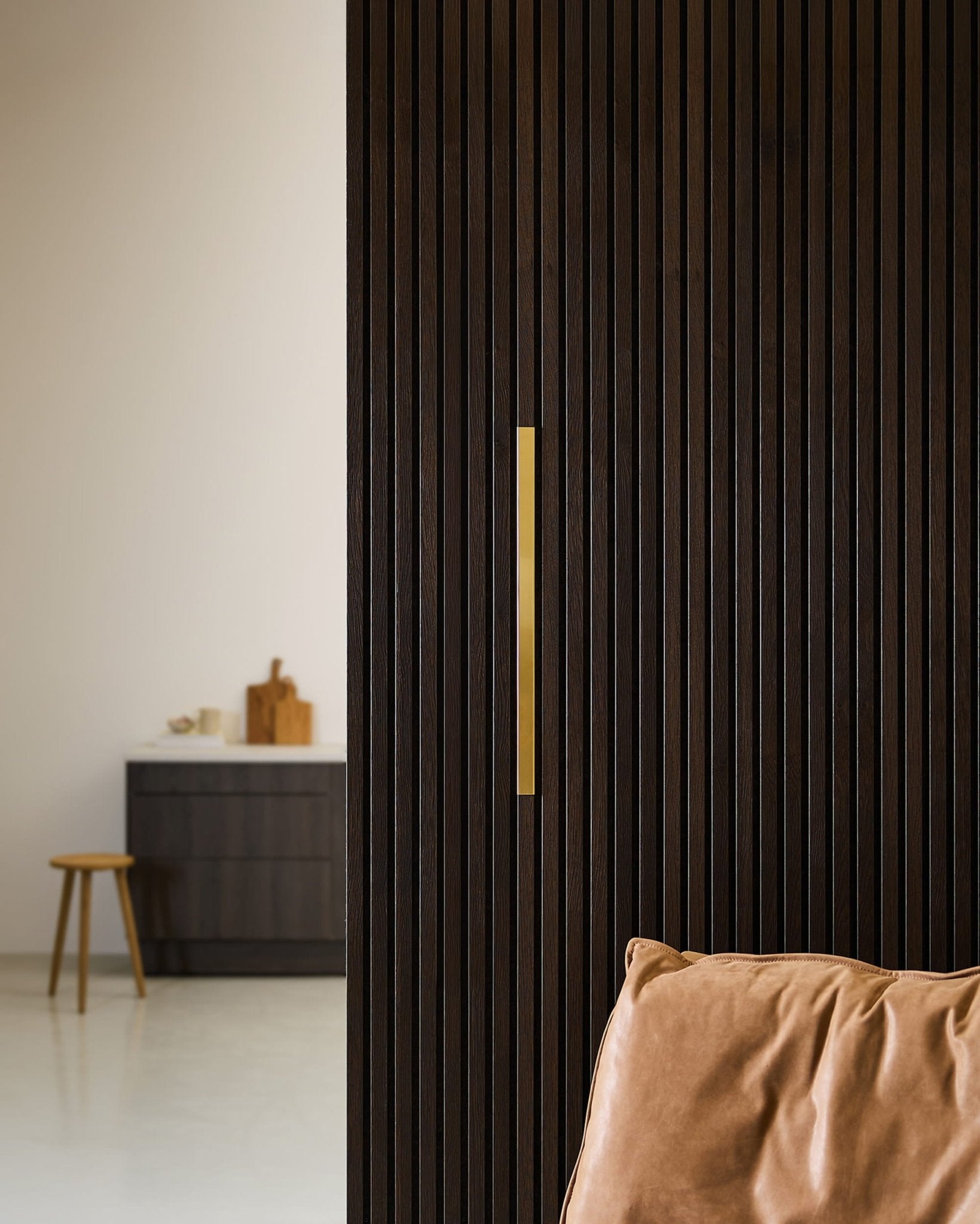 Minimalist room featuring a WoodUpp Akupanel Liva | Cover / Brass, 60 cm, next to a leather chair, stool, and dark cabinet in a neutral setting.