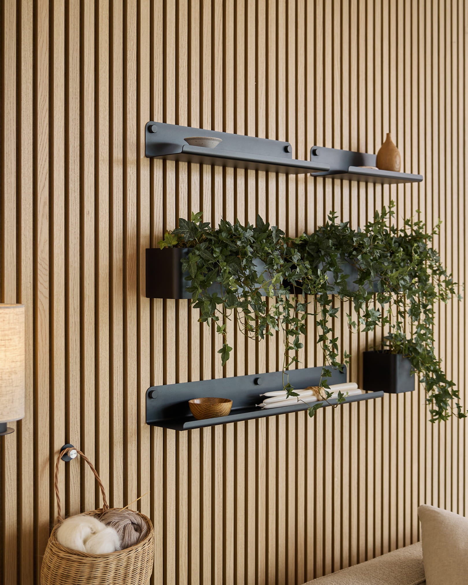 Three black Norr | Plant Box shelves by WoodUpp with decor and a plant are mounted above a wicker basket of yarn, set against a green acoustic slatwall panel.