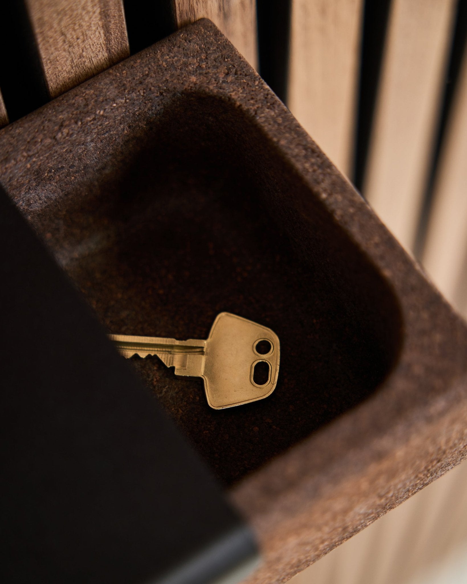 A brass key rests in a Norr Key Tray/Smoked Cork, 35.9 cm by WoodUpp, placed on a brown container in front of an Akupanel acoustic wood panel background.