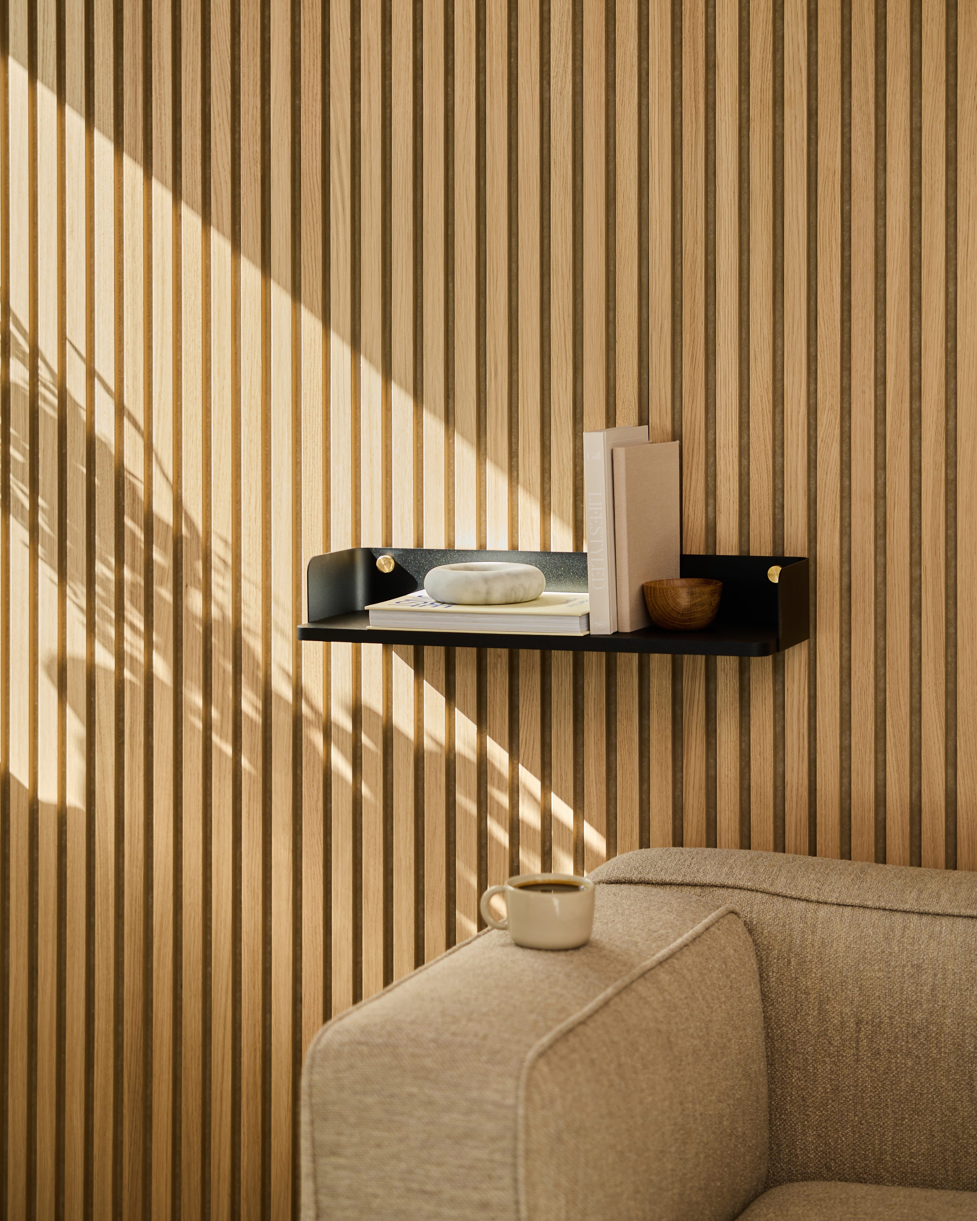 A Linn | Bookshelf / Black Oak by WoodUpp displays decor above a beige sofa. Sunlight casts shadows on the striped acoustic wood panel wall.