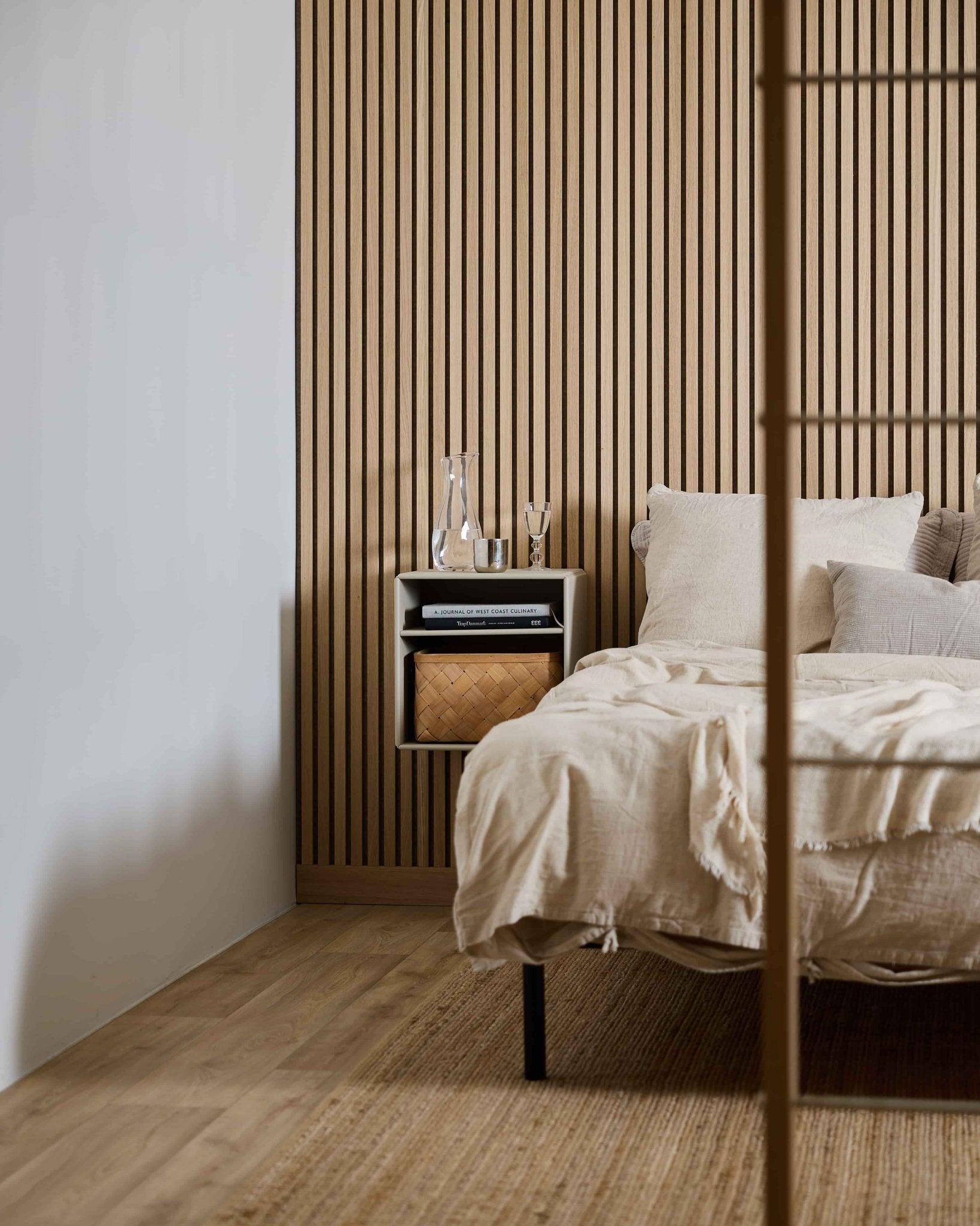 Minimalist bedroom: WoodUpp Arka Bookcase, beige bedding, Akupanel wood slat wall, floating nightstand, glass carafe, woven basket, light floor.