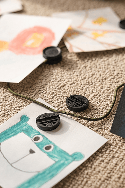 Children’s drawings hang from the WoodUpp Vindar | Gallery String with neodymium clips; green bear artwork rests on beige carpet below the acoustic panel.