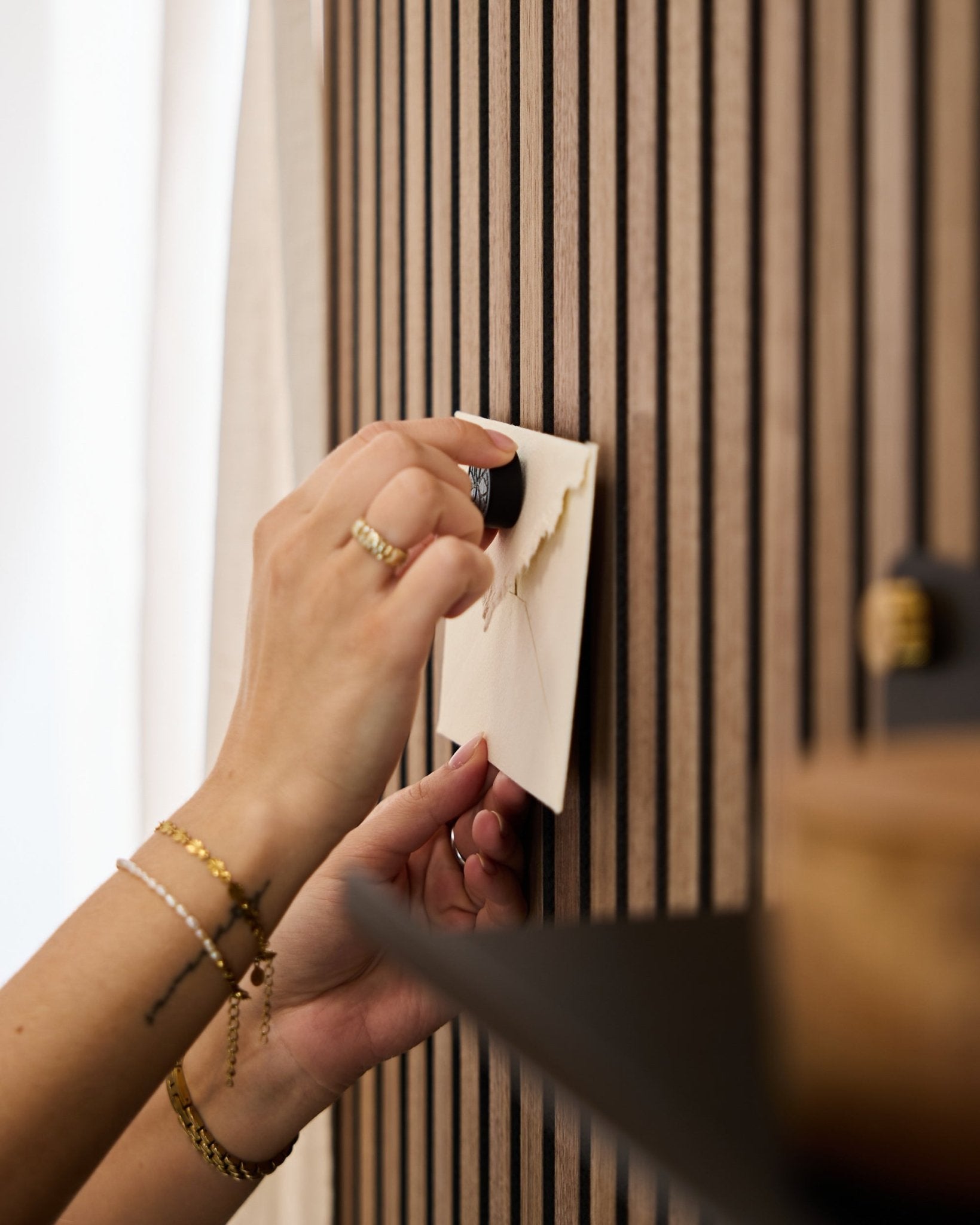 A person wearing gold bracelets and rings attaches an envelope to a WoodUpp oak acoustic slatwall panel using the Norr Poster Magnet set in Black Oak.
