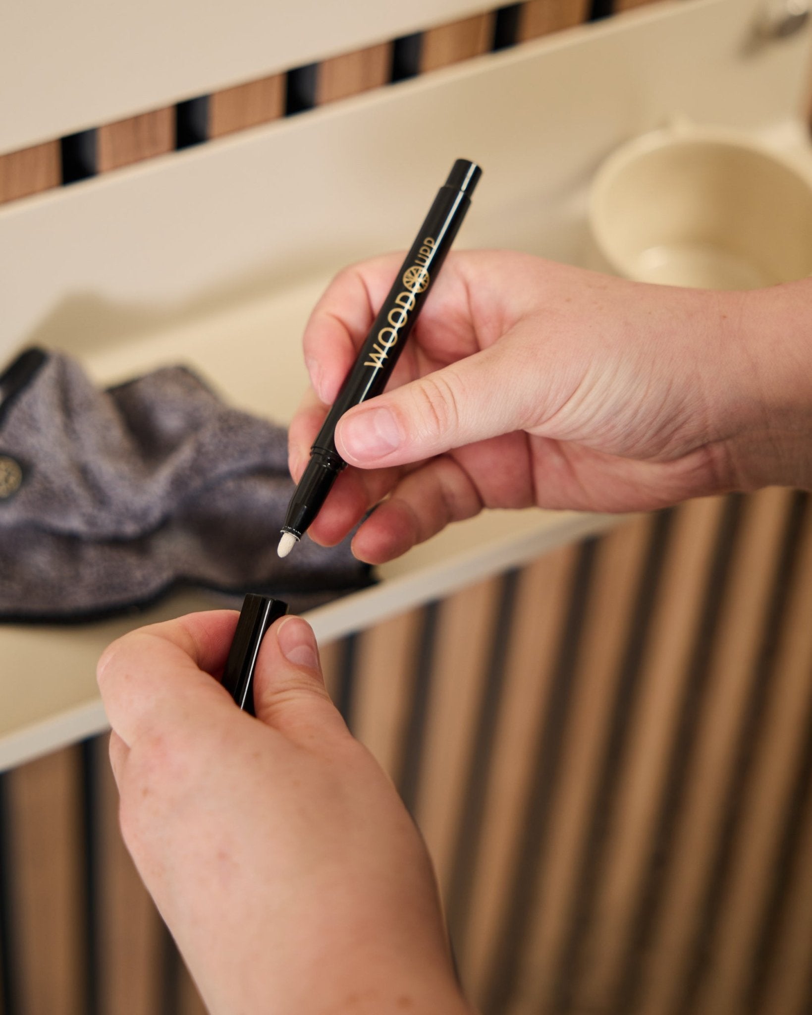 A person holds an open WoodUpp Norr | Blackboard set marker beside a microfiber cloth and a beige cup, products designed for use with acoustic wood panels.