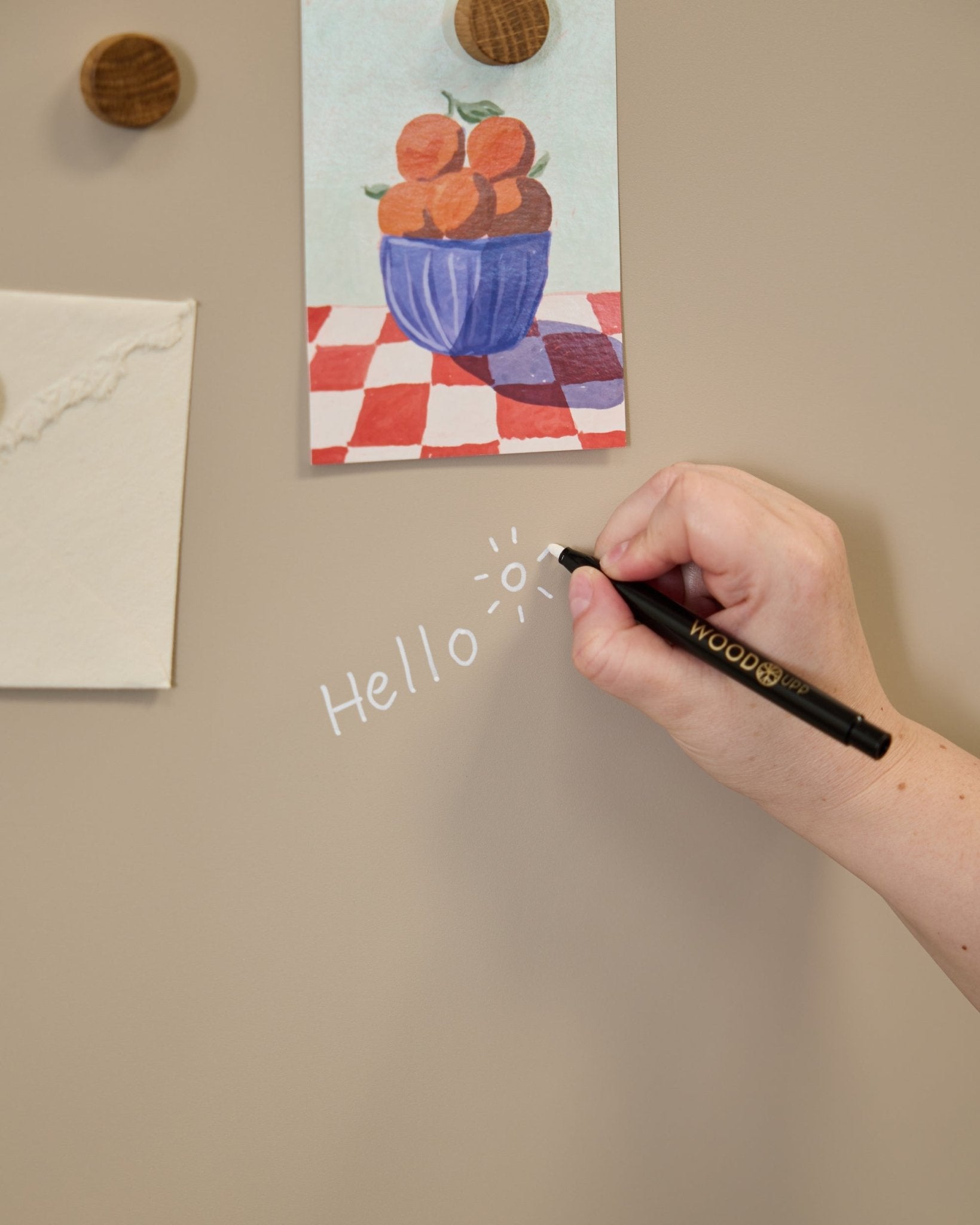 A hand writes hello with a sun doodle on the WoodUpp Norr Blackboard, set beside art attached with round magnets on an acoustic panel.