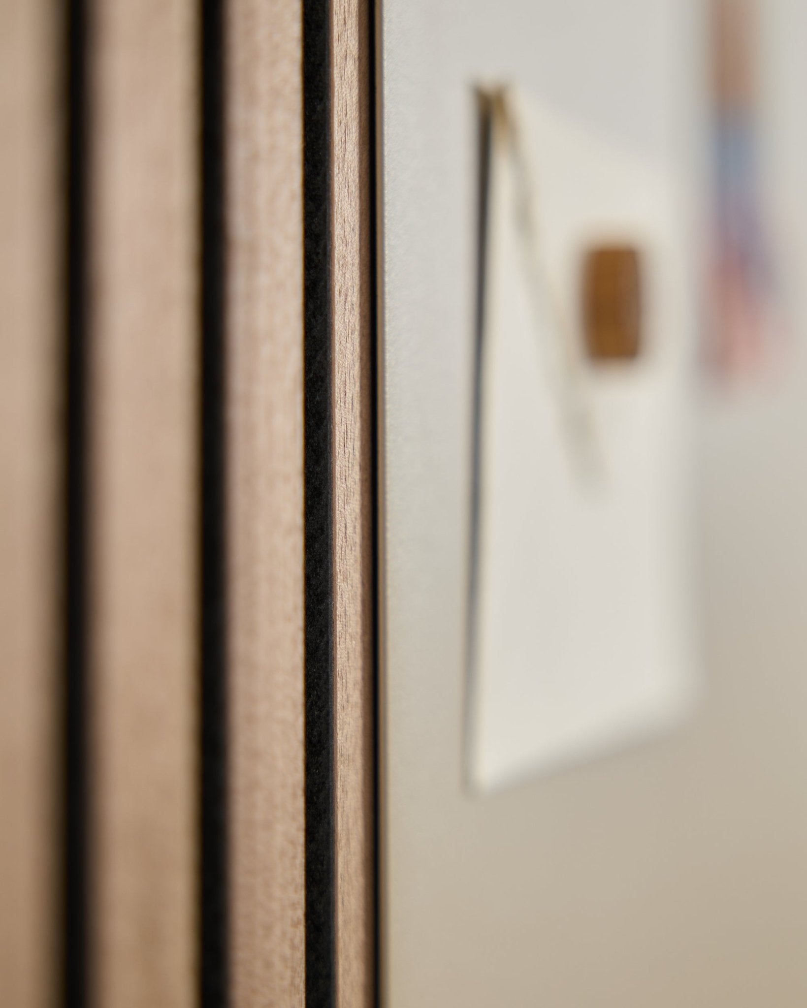 Close-up of a WoodUpp Akupanel acoustic wood panel with vertical slats, next to a blurred large beige Norr Magnetic Board in the background.
