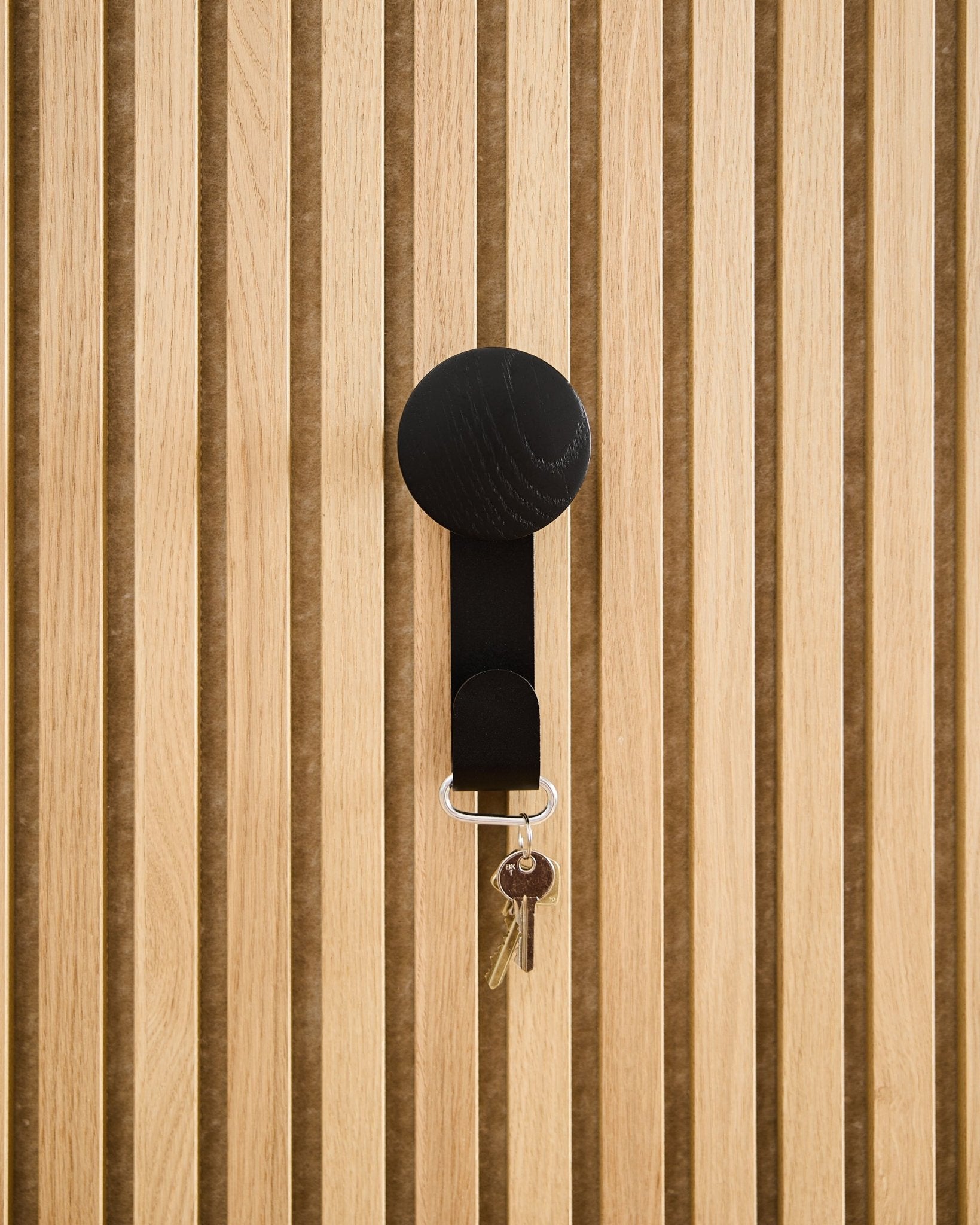 A Linn Vertical Hanger in Black Oak by WoodUpp holds a keyring with two keys and a round tag, mounted on a textured Akupanel acoustic wood panel.