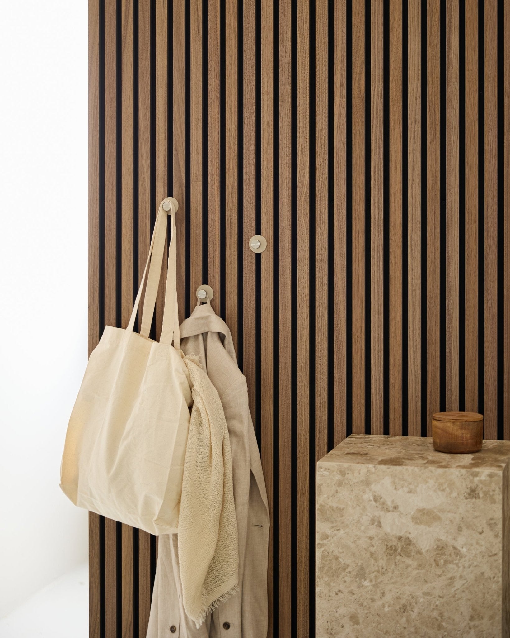 A beige tote bag and a light fabric hang from a WoodUpp Norr Coat Hook (Beige/Stainless Steel) mounted on a vertical Akupanel acoustic panel beside a stone surface.