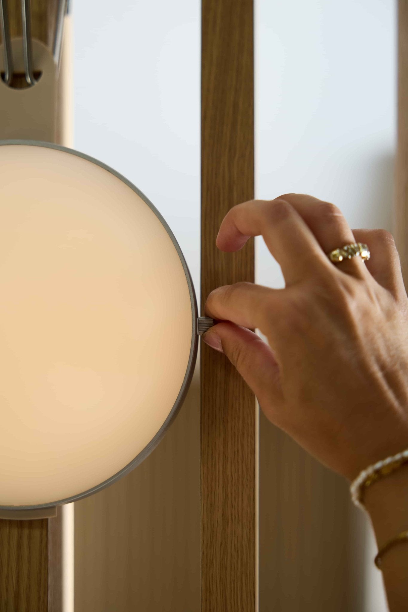 A hand with a ring adjusts a Vala | Portable LED Lamp by WoodUpp, mounted on a light wood Akupanel acoustic slatwall panel.