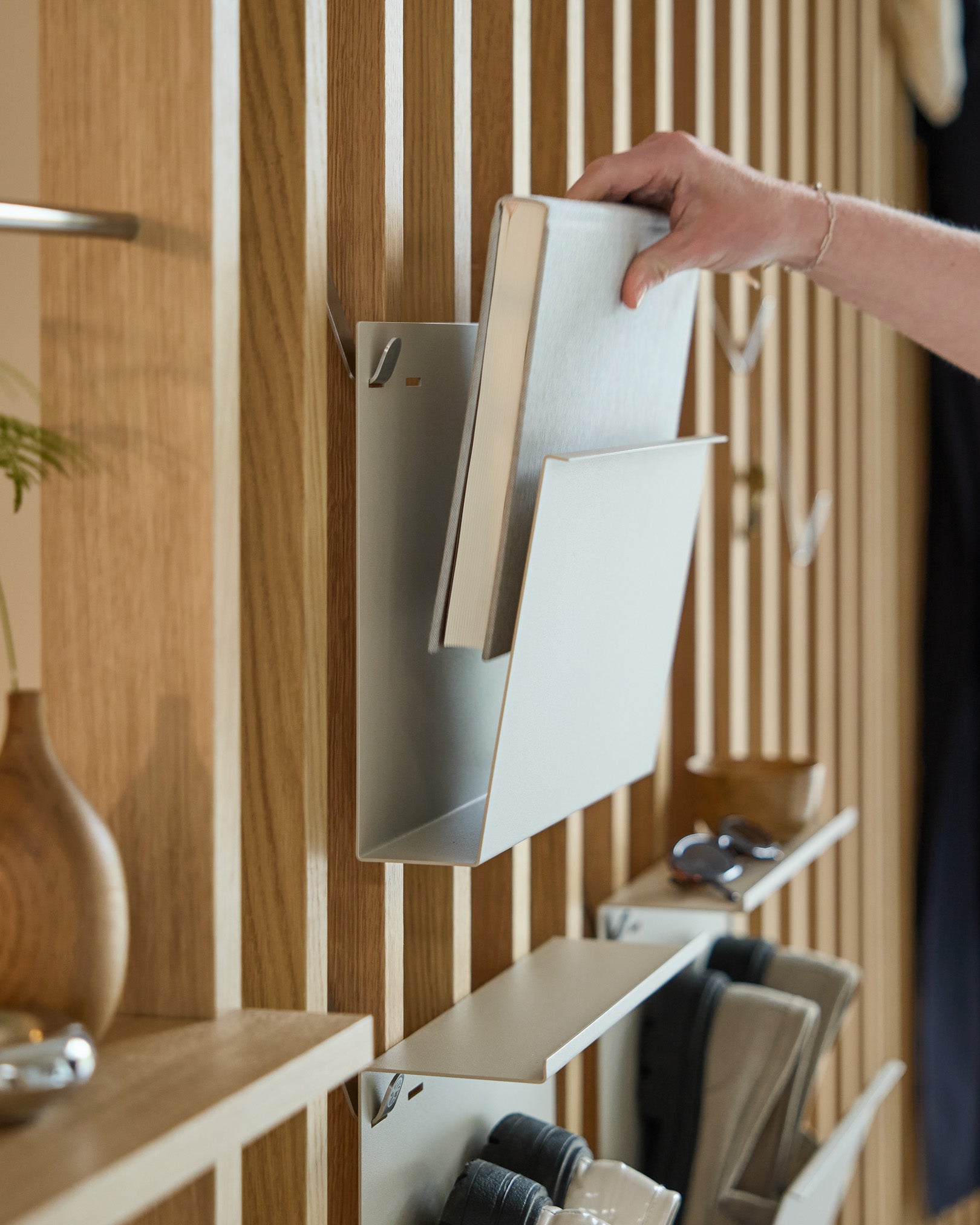 A hand places Nell | Magazine Storage / Beige by WoodUpp onto a powder-coated steel shelf mounted on a Nordic wooden acoustic panel with nearby items.
