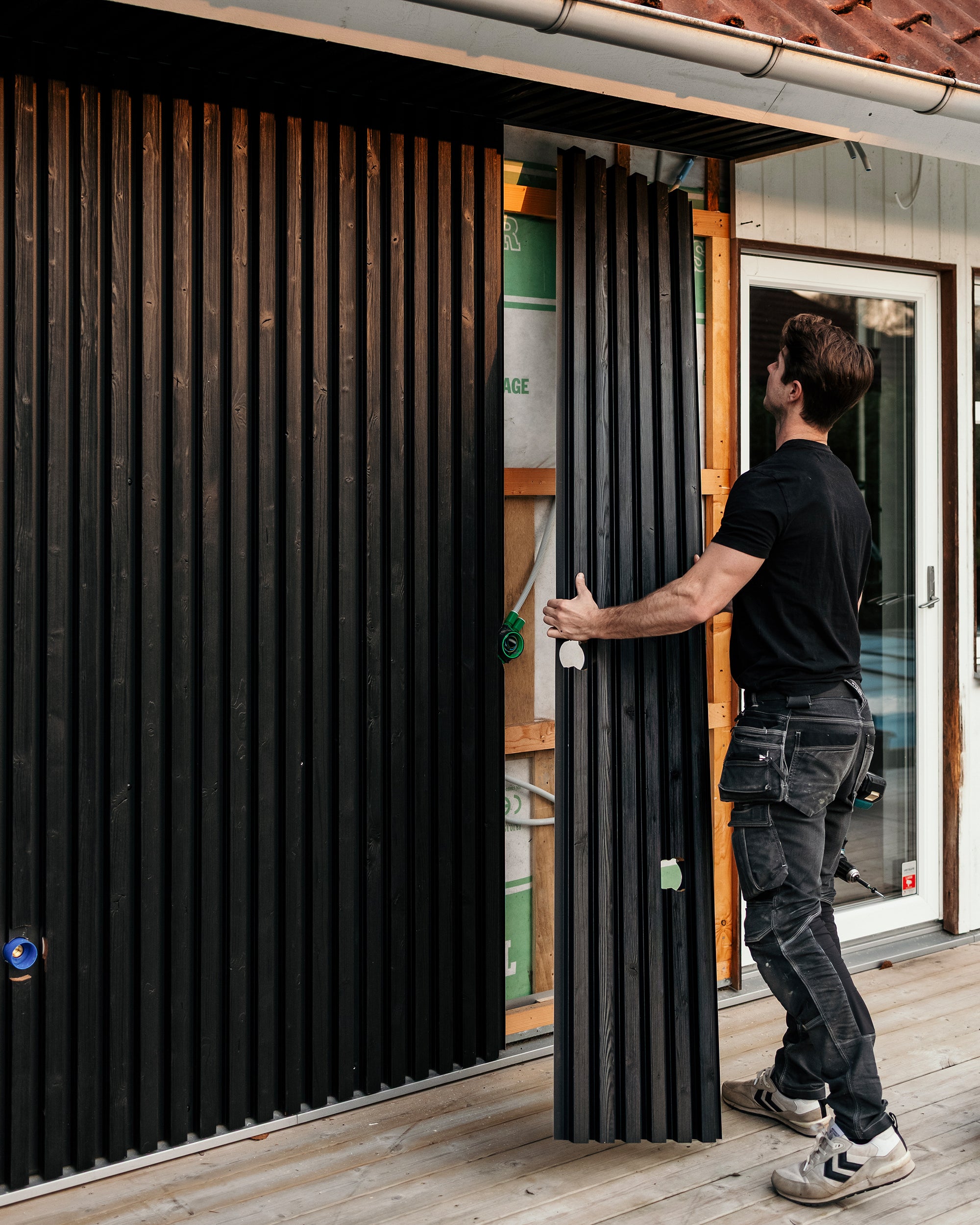 A man installs vertical black Aluwood facade panels on a homes exterior wall while standing on a newly built deck.