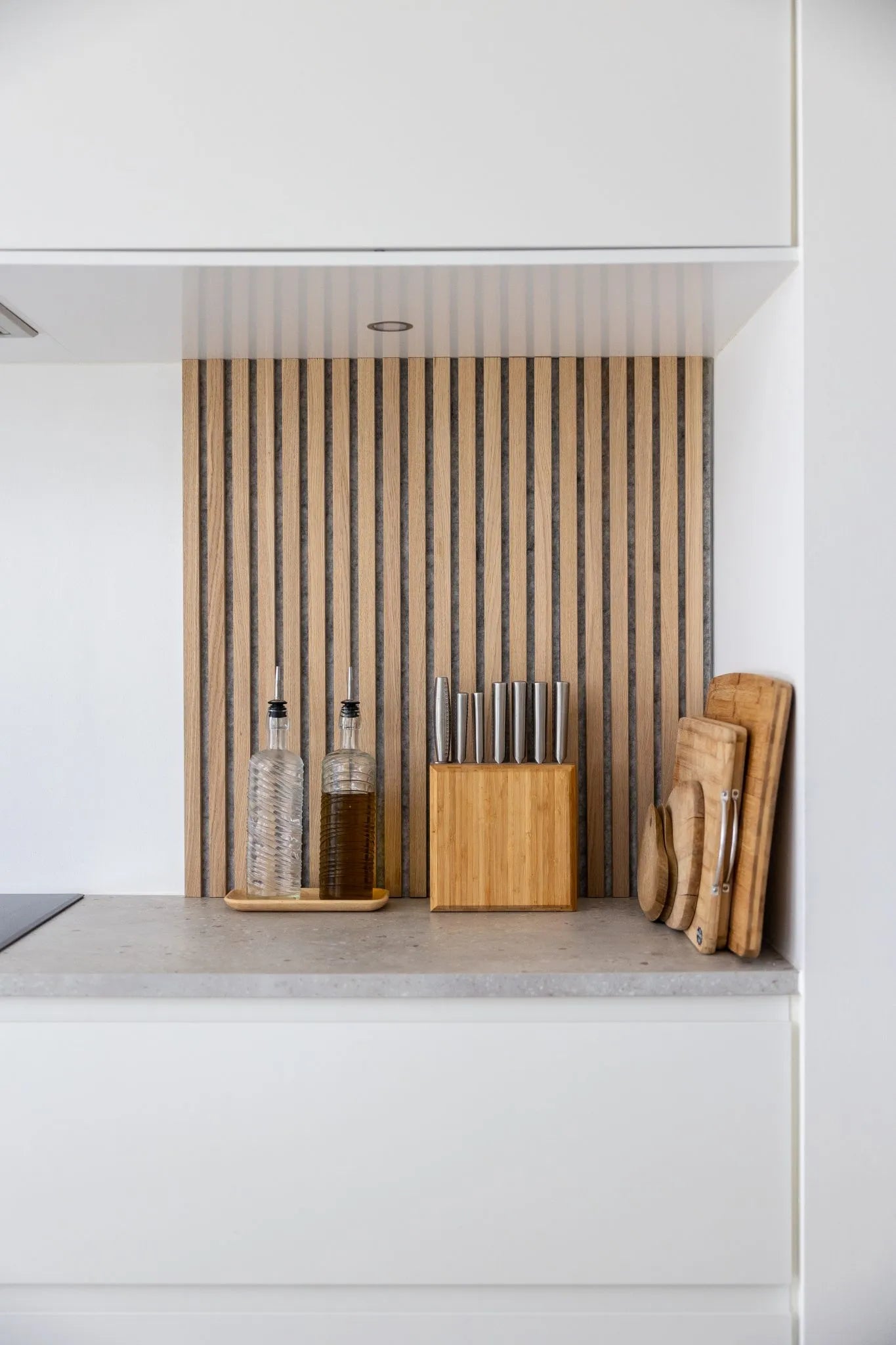 Modern kitchen counter with oil bottles, wooden knife block, and stacked cutting boards set against a Nordic Akupanel acoustic wood slat backsplash and white cabinets.