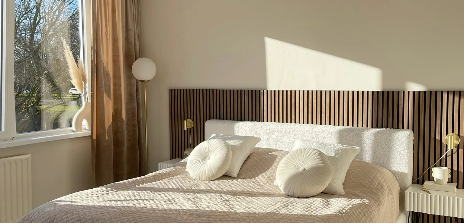Sunlit modern bedroom with a beige quilted bed, round pillows, Akupanel wood slat headboard, and minimal decor beside a large window with sheer curtains.