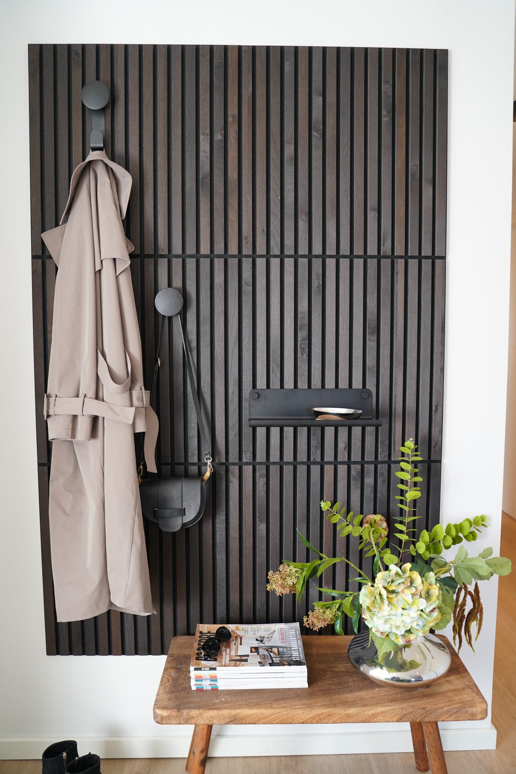 Nordic acoustic slatwall panel with coat hooks holding a beige trench coat and black bag; bench in front displays books, sunglasses, and a vase of flowers.