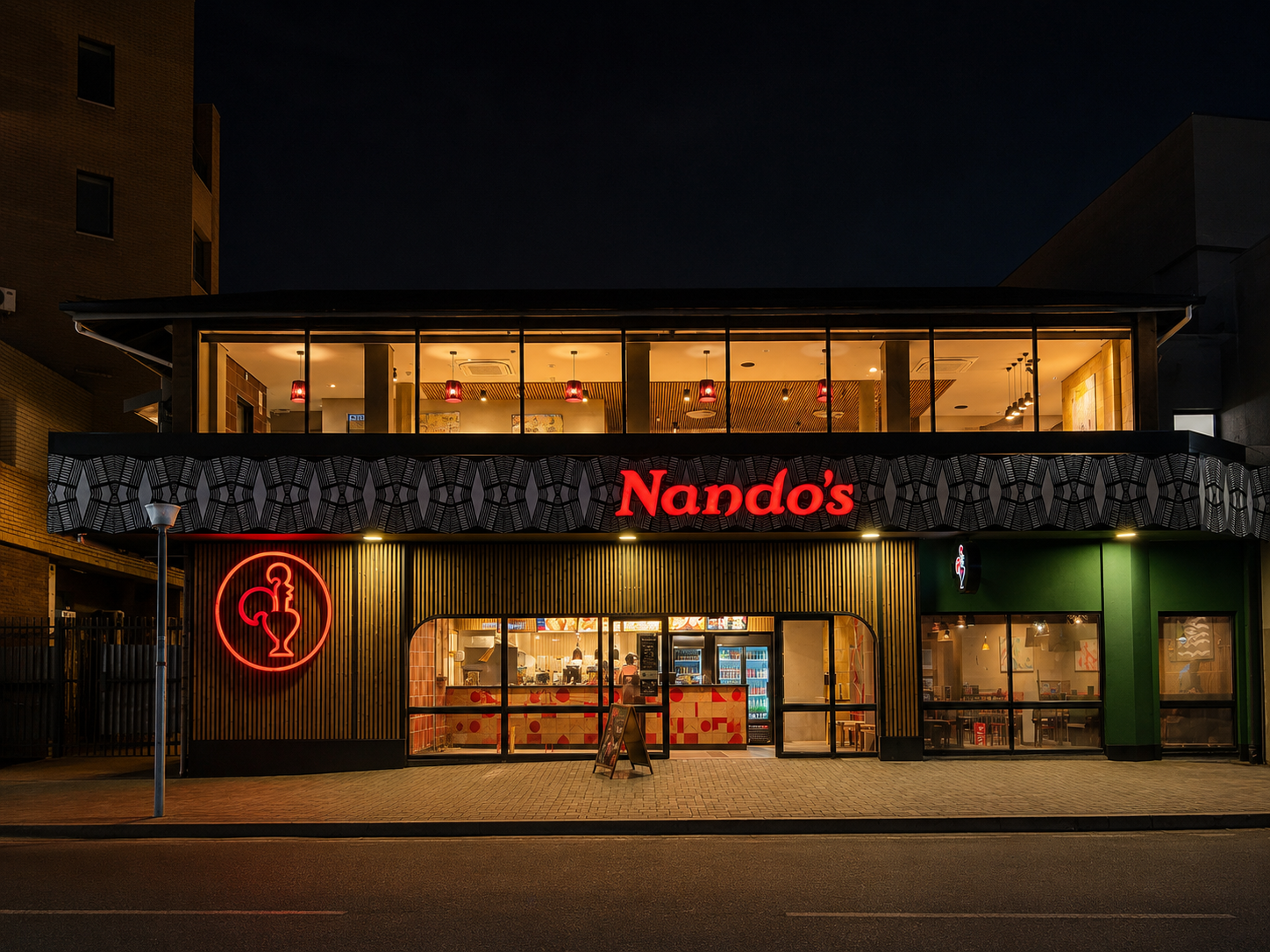 Nandos restaurant at night with bright red neon signage and Peri-Peri chicken logo; interior lighting reveals tables, chairs, and acoustic panels inside.