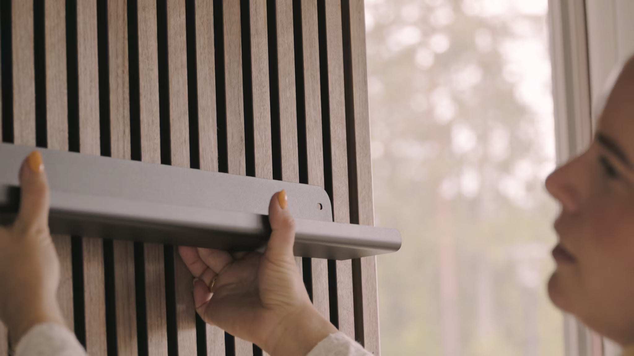 A person installs a gray shelf onto a Nordic Akupanel acoustic wood slat wall beside a window looking out to trees.