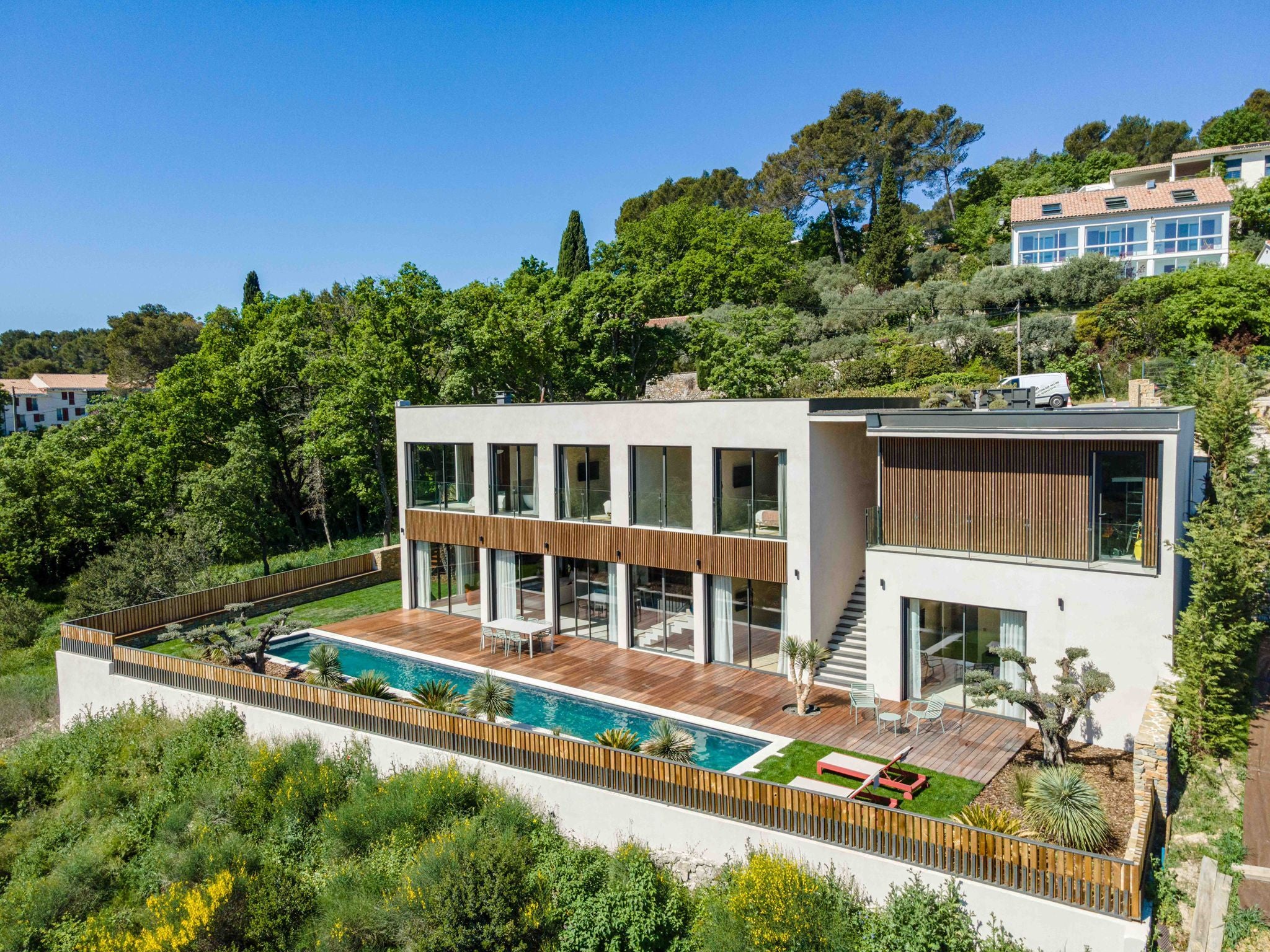 Modern two-story smart home with large windows, wooden deck, and long pool, featuring Nordic facade panels, set among greenery and hills under a blue sky.