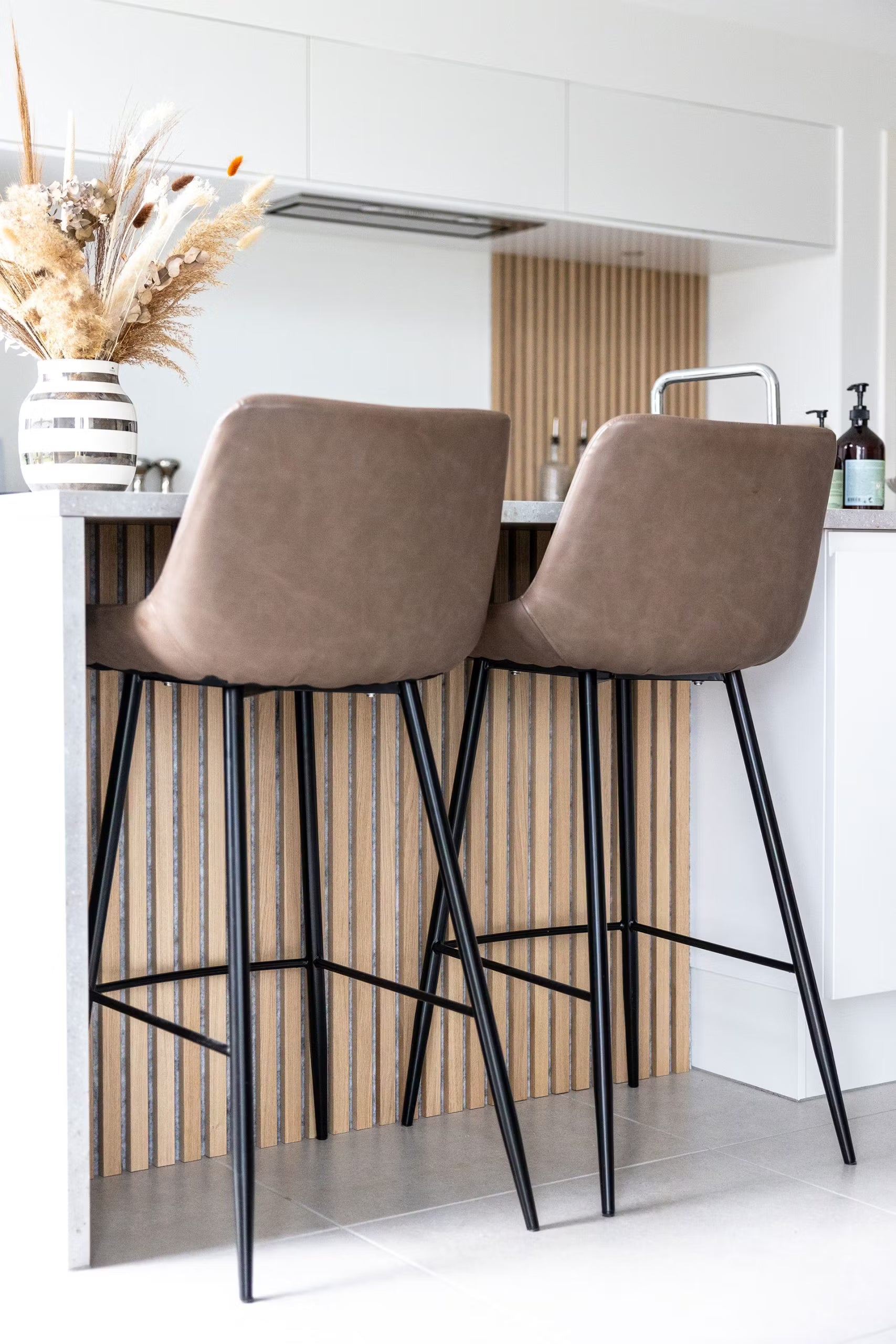 Brown bar stools with black legs at a kitchen island, featuring a Nordic acoustic wood slatwall panel and a vase with dried flowers on the countertop.