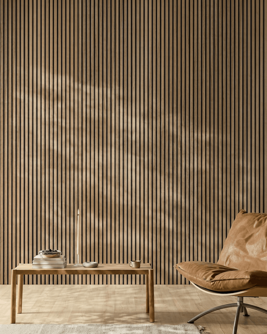 Minimalist room with a WoodUpp Akupanel acoustic panel (English Walnut slats, black felt), tan leather chair, light wood table, candle, books, cup, white rug in sunlight.