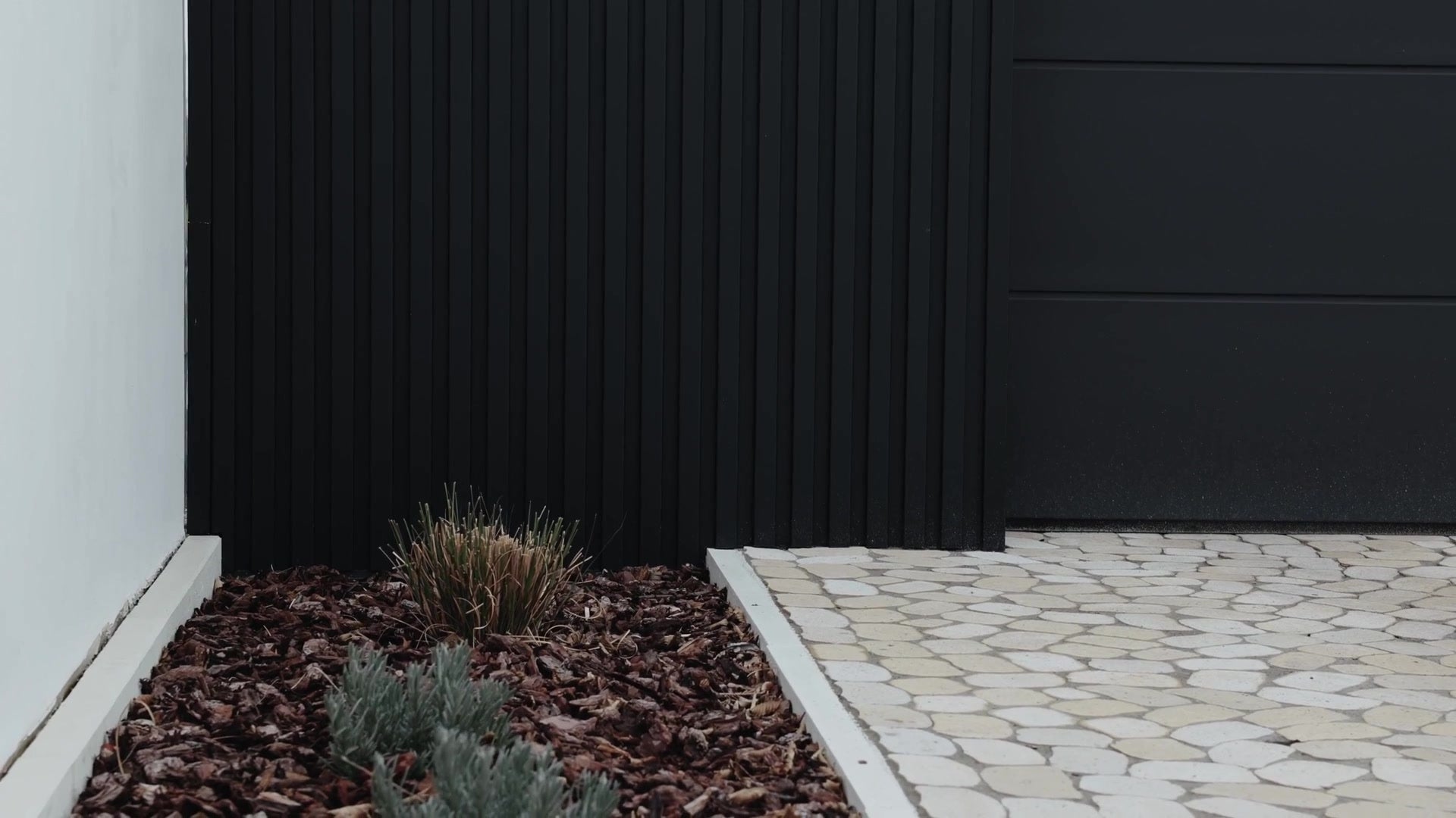 Black fluted garage wall with a garage door, a gravel and bark planting bed and a white house wall