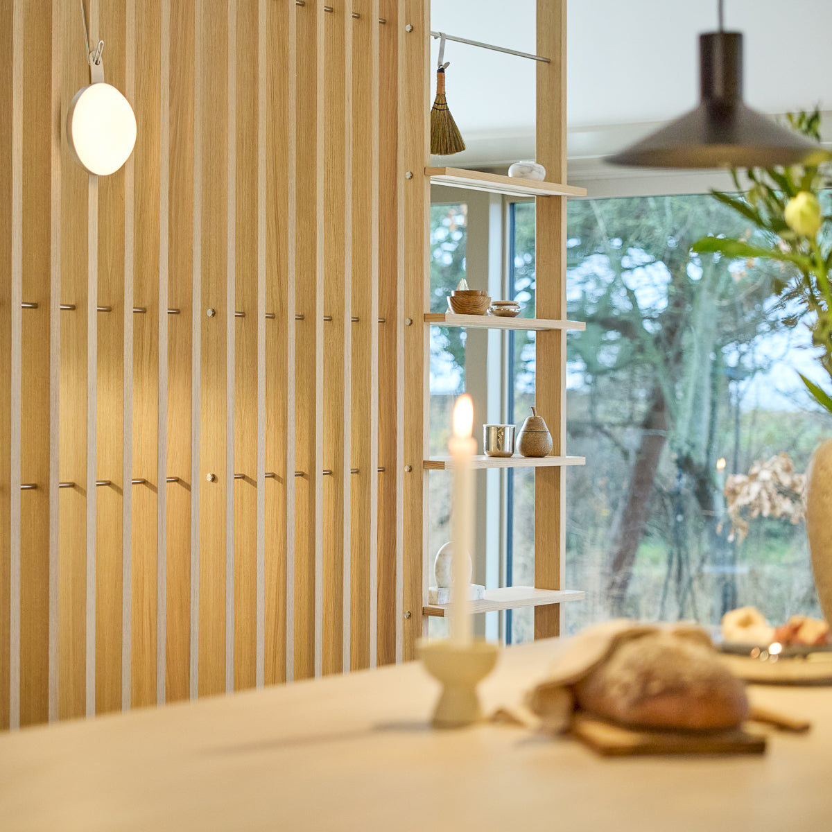 Minimalist interior with a Nordic EMBRACE wooden room divider, lit candle, fresh bread on table, curated decor on shelves, and window framing trees outside.