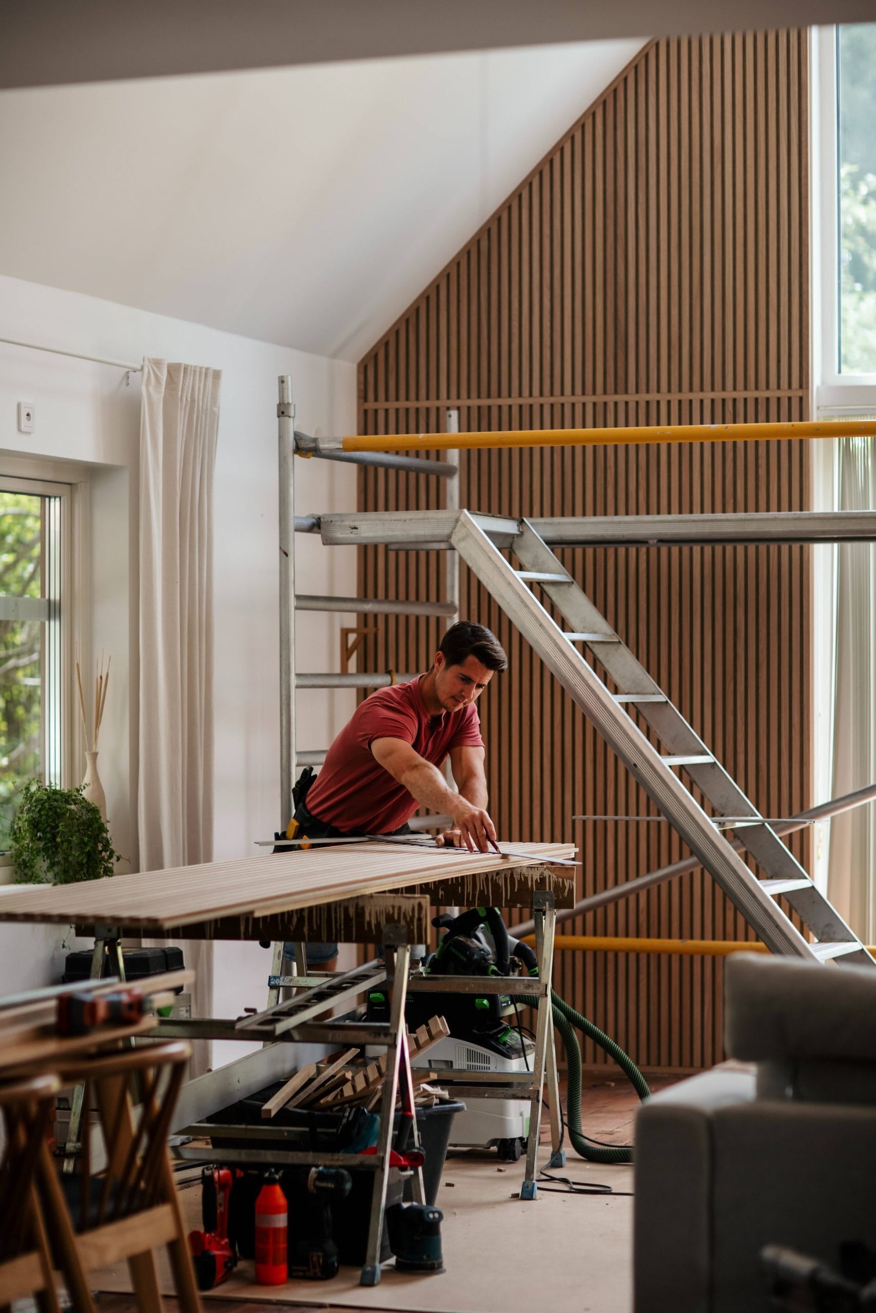 A man measures a wooden plank on a workbench in a modern, partially renovated room with scaffolding and large windows, suggesting acoustic panel installation.