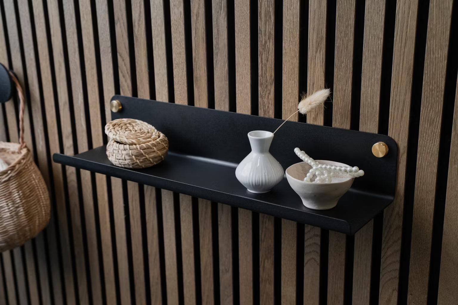 A black wall shelf with a small wicker basket, white vase, and ceramic bowl of pearls mounted on a Nordic wooden acoustic slatwall panel.