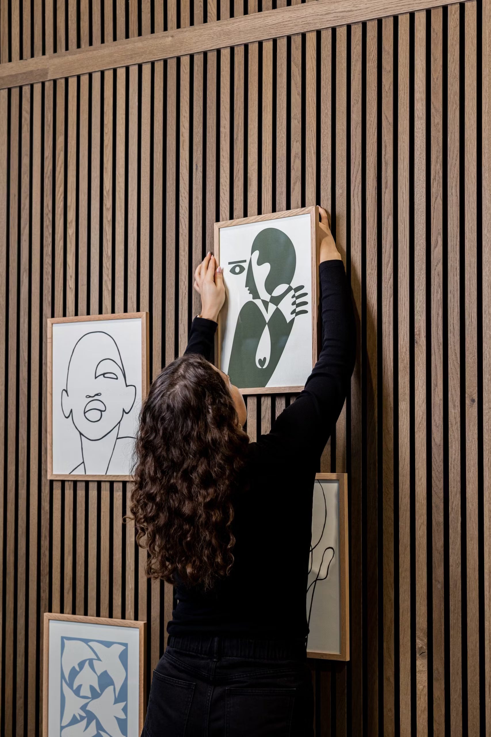 A person with curly hair hangs a WoodUpp® CREATE DUPPs accessory on a wooden acoustic slatwall panel, surrounded by minimalist art and modern decor.